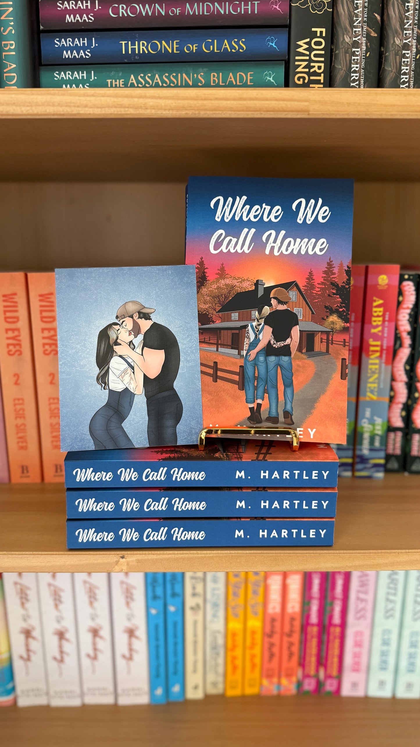 Bookshelf with multiple copies of 'Where We Call Home' by M. Hartley and other books.