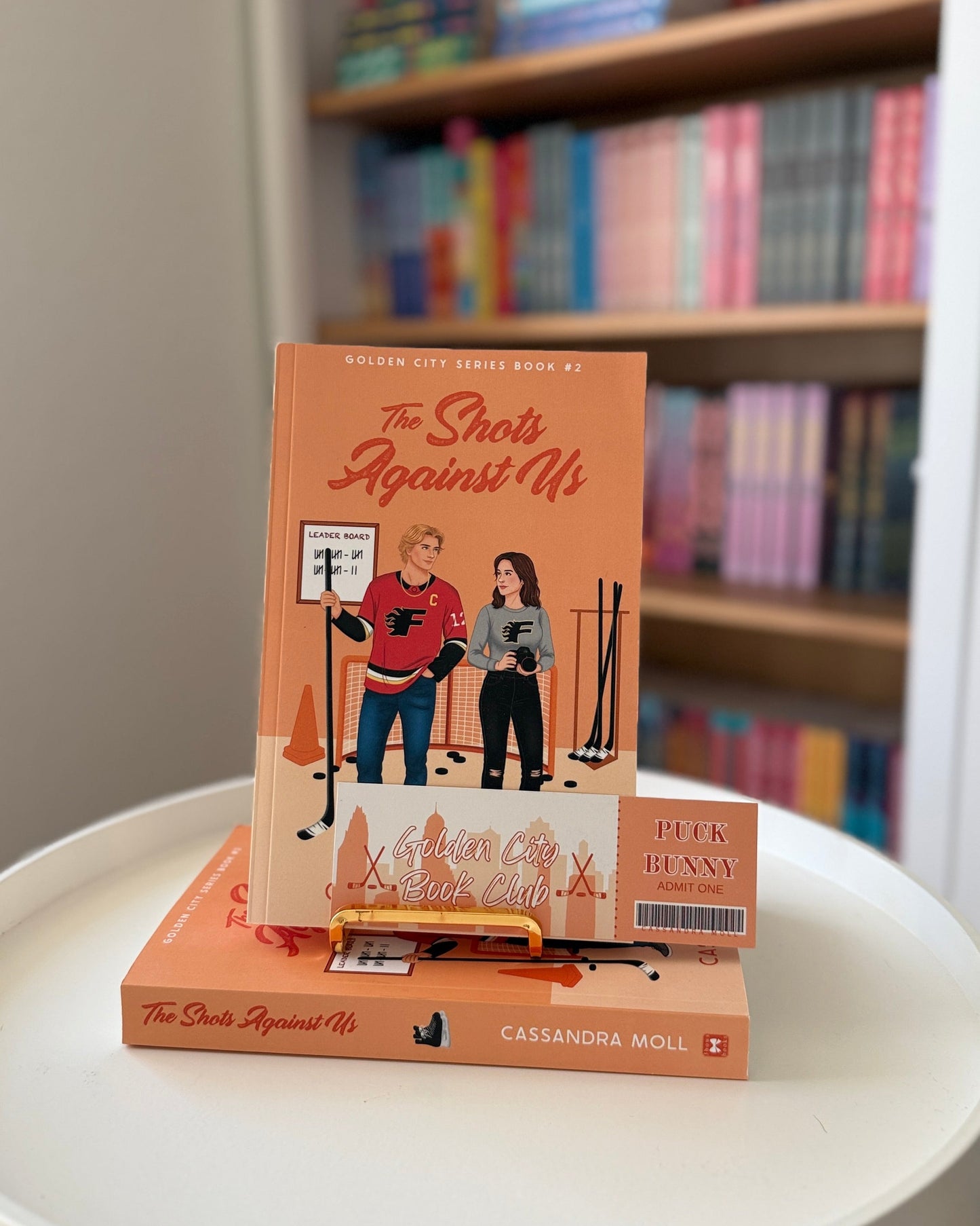 Book titled 'The Shout Against Us' on a white surface with a bookshelf in the background.