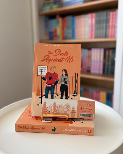 Book titled 'The Shout Against Us' on a white surface with a bookshelf in the background.