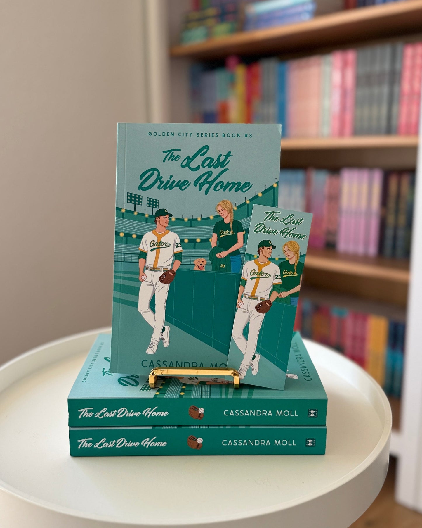 Stack of books titled 'The Last Drive Home' on a white stool with a bookshelf in the background.