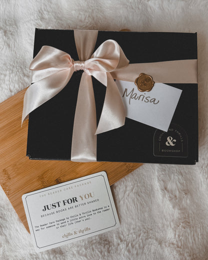 A black gift box tied with a pale pink satin ribbon and bow, placed on a wooden board over a white faux-fur surface. A small card reading 'Marisa' and a separate 'Just for You' card are arranged nearby.