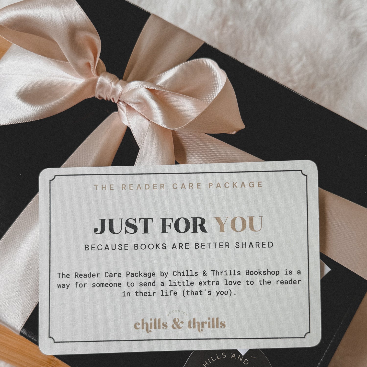 A black gift box tied with a pale blush satin ribbon, opened to reveal a white card that reads 'Just for You' and 'The Reader Care Package by Chills & Thrills Bookshop.' A round black sticker on the box shows the Chills & Thrills Bookshop logo.