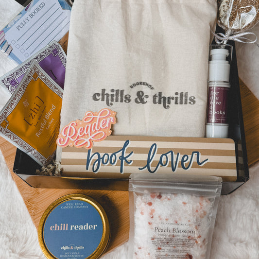 A book lover gift box set on a white faux fur surface, featuring a cream drawstring bag with 'chills & thrills' printed on it, a small bottle, a tin labeled 'chill reader', a bag of peach blossom salt, and other cozy reading accessories.