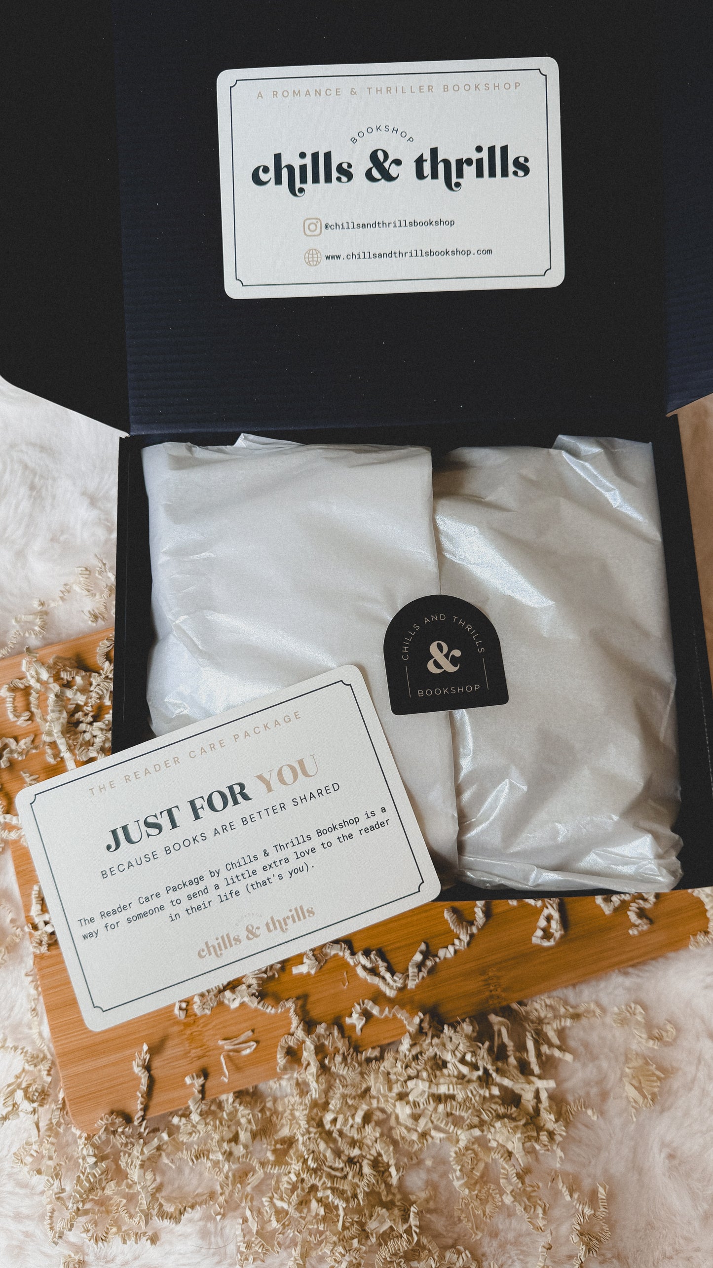 An open black gift box from chills & thrills containing two white-wrapped packaged items, with printed cards placed in front. The box is styled on a wooden board with shredded paper filler over a white textured surface.