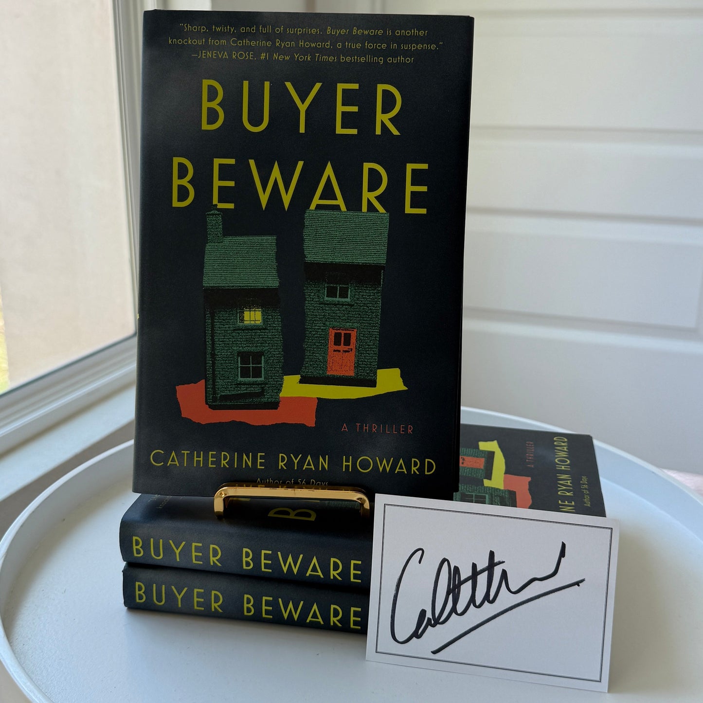 Book 'Buyer Beware' by Catherine Ryan Howard displayed on a white round table next to a white card with a black signature, with a window and paneled wall in the background.