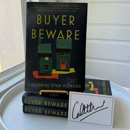 Book 'Buyer Beware' by Catherine Ryan Howard displayed on a white round table next to a white card with a black signature, with a window and paneled wall in the background.