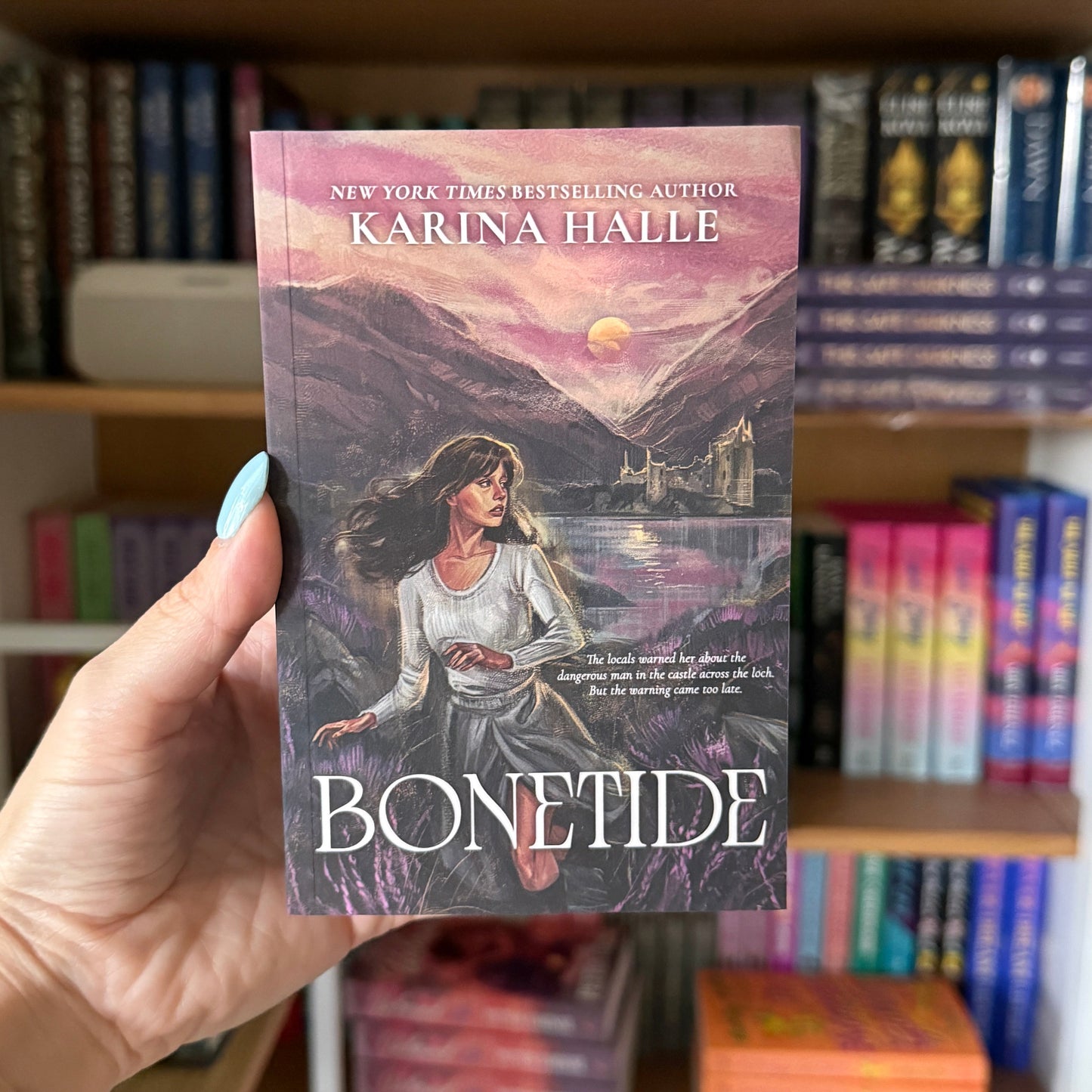 A hand holds a copy of the book 'Bonetide' by Karina Halle in front of a blurred bookshelf.