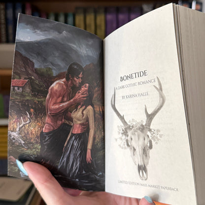 A hand holds an open book in a library or bookstore, showing the illustrated front cover and the title page with the text 'BONETIDE' and a deer skull illustration. Bookshelves with books are visible in the blurred background.