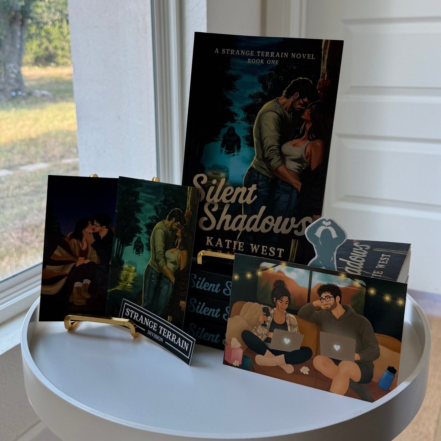 A book and its promotional cards and cover art are arranged on a small round white table in a bright room near a window. The main item is the book titled "Silent Shadows" by Katie West, with additional printed materials in front.