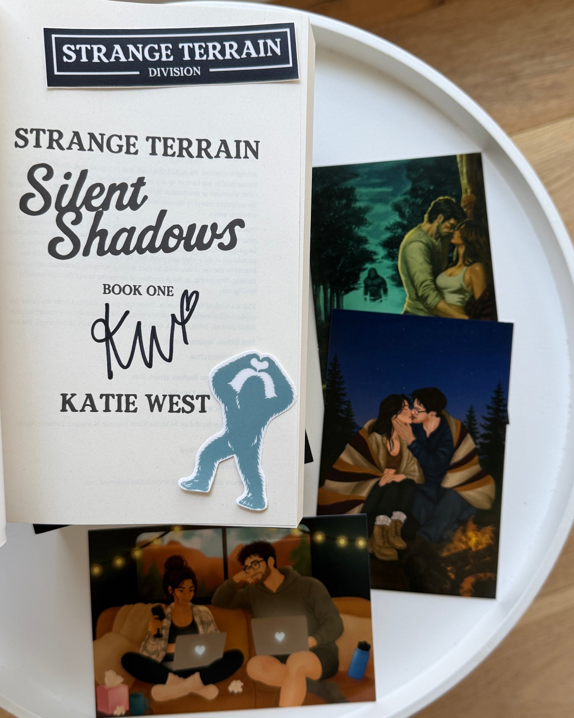 A book titled 'Strange Terrain Silent Shadows' by Katie West is displayed on a white round tray, alongside printed character art cards. The book cover features a white background with black and gray text and a blue silhouette graphic.