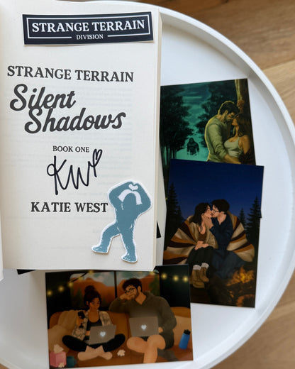A book titled 'Strange Terrain Silent Shadows' by Katie West is displayed on a white round tray, alongside printed character art cards. The book cover features a white background with black and gray text and a blue silhouette graphic.