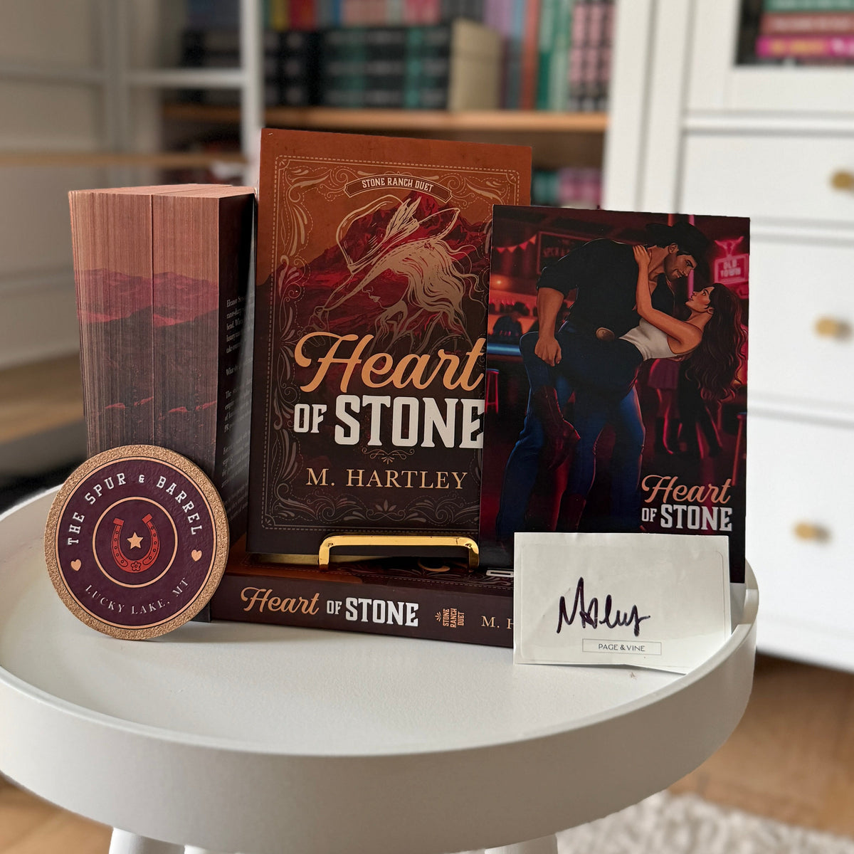 A book-themed gift set on a white round table, featuring the book 'Heart of Stone' by M. Hartley, a matching book or box, a small round wooden plaque, and a white card with 'Maeve' written on it.