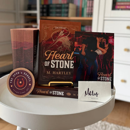A book-themed gift set on a white round table, featuring the book 'Heart of Stone' by M. Hartley, a matching book or box, a small round wooden plaque, and a white card with 'Maeve' written on it.