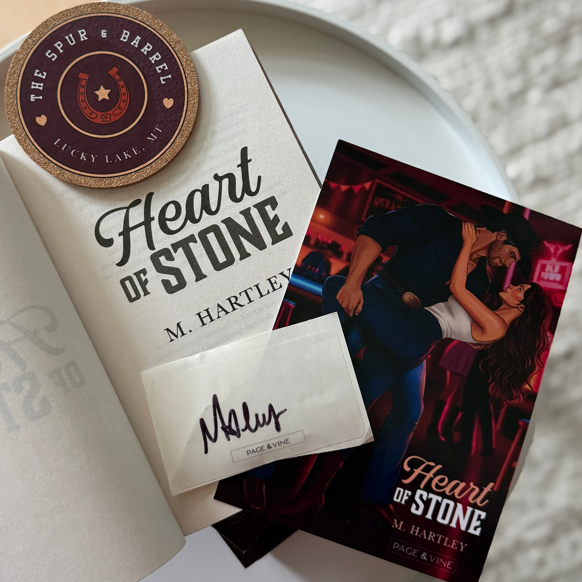 A styled flat-lay of a book and its packaging on a white plate, set on a light rug over a wooden floor. The book is titled 'Heart of Stone' by M.