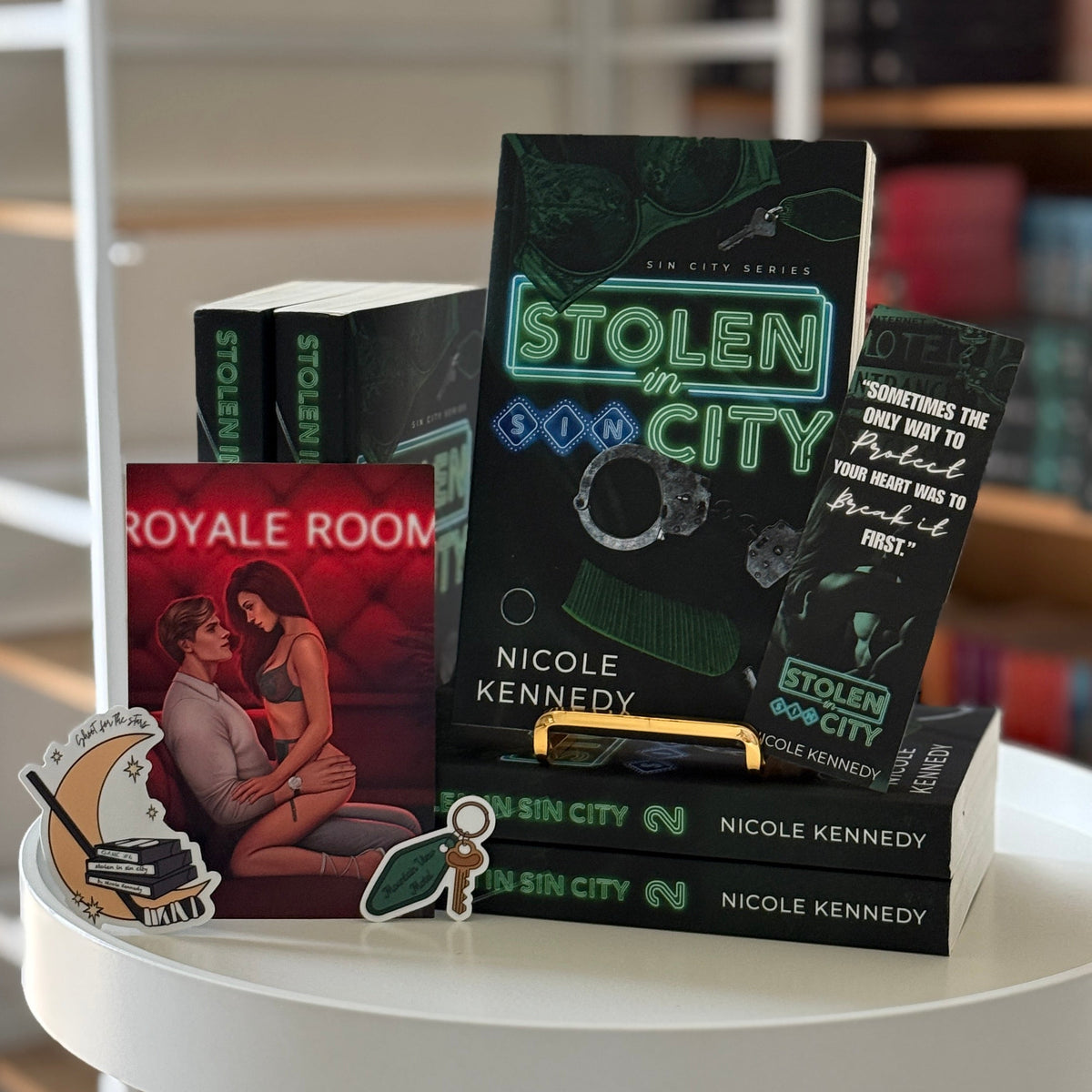 A stack of Nicole Kennedy books, including 'Stolen City' and 'Royal Roon', displayed on a small white round table in a book-filled room.