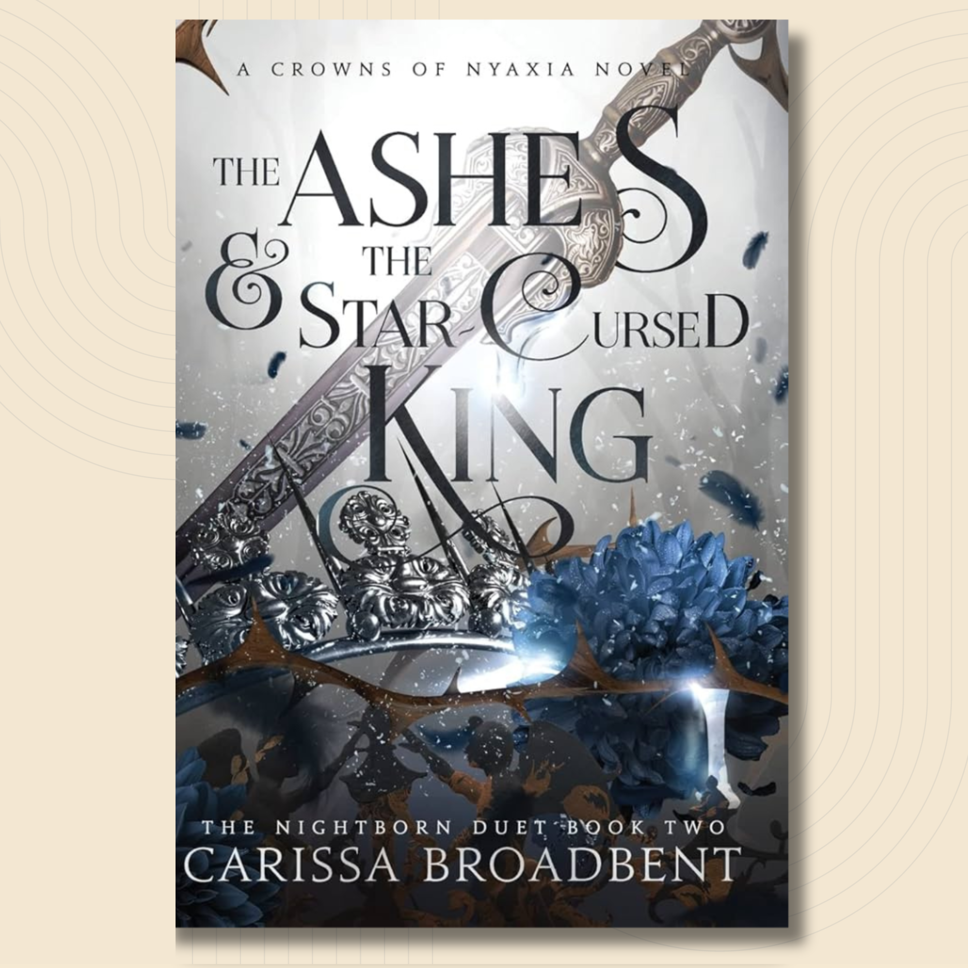 Book cover for 'The Ashes & the Star Cursed King' by Carissa Broadbent, featuring a silver ornate sword, blue flowers, and dark gothic typography on a light background, shown against a beige textured backdrop.