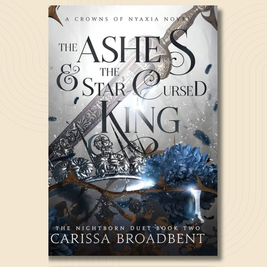 Book cover for 'The Ashes & the Star Cursed King' by Carissa Broadbent, featuring a silver ornate sword, blue flowers, and dark gothic typography on a light background, shown against a beige textured backdrop.