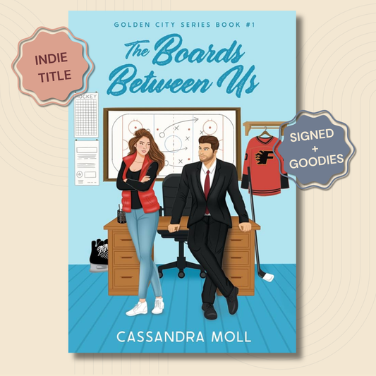 Book cover for 'The Boards Between Us' by Cassandra Moll, featuring an illustrated office scene with a woman in a red-and-black top and jeans, a man in a black suit, a sports jersey, and a whiteboard. The cover is shown on a beige background with 'INDIE TITLE' and 'SIGNED + GOODIES' badges.