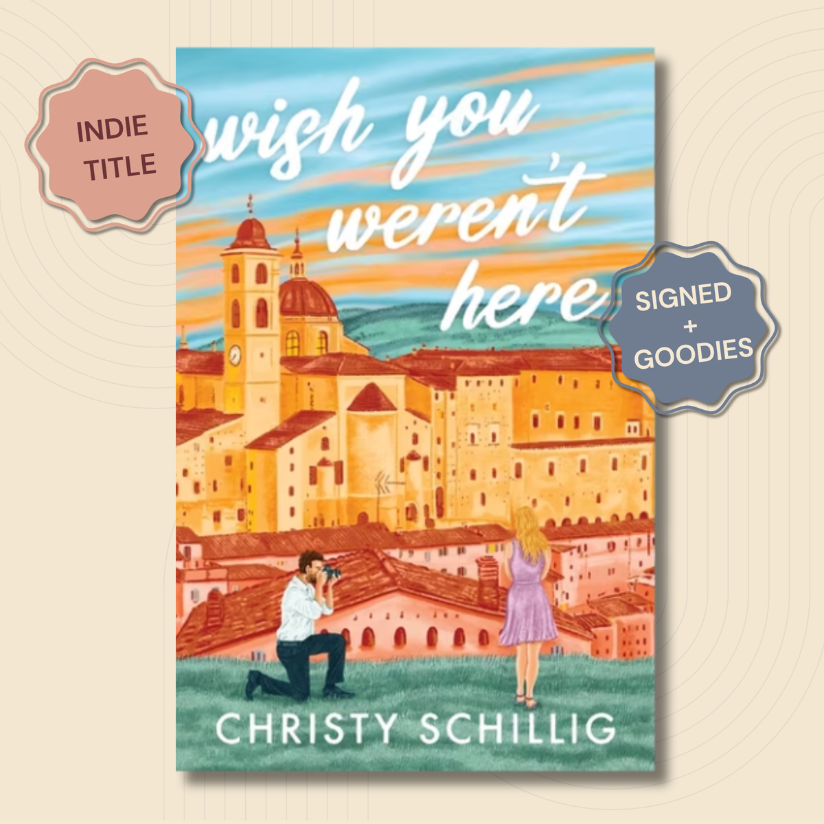 Book cover for 'wish you weren't here' by Christy Schillig, featuring an illustrated cityscape with warm orange buildings, a blue sky, and two small figures in the foreground. Two decorative badges reading 'INDIE TITLE' and 'SIGNED + GOODIES' are overlaid on the image.
