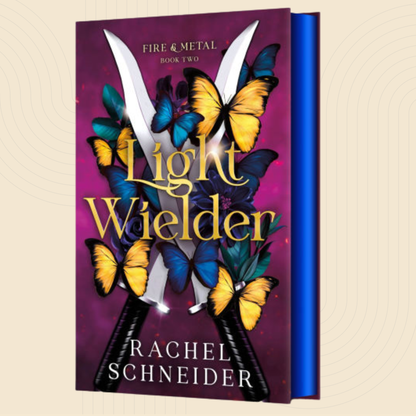 Book cover for 'Light Wielder' by Rachel Schneider, showing a purple background with large gold title text, blue and yellow butterflies, and crossed metallic shears. The book is shown standing upright with a blue spine on a light beige background.
