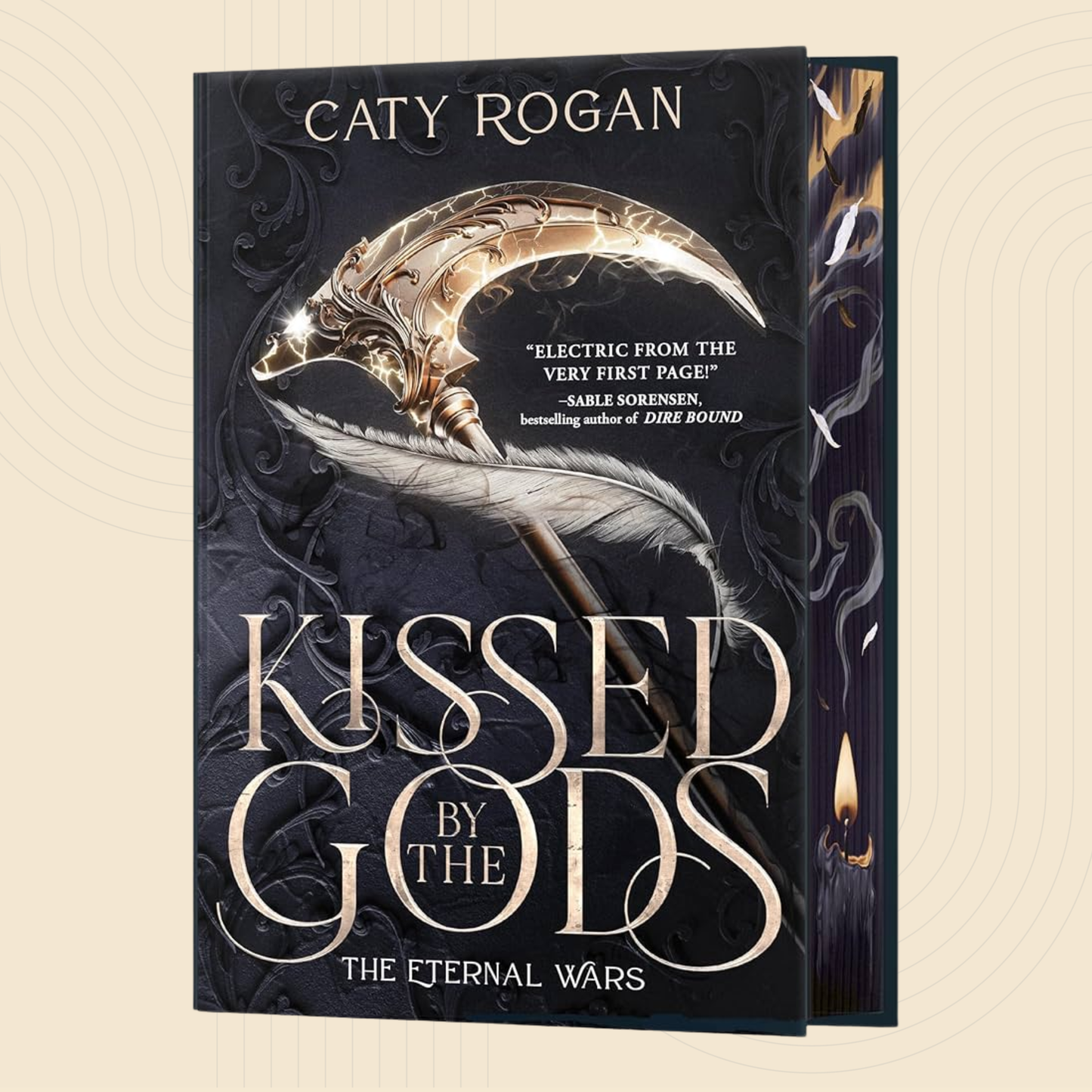 Book cover for 'Kissed by the Gods' by Caty Rogan, featuring a large ornate gold crescent moon and feather illustration on a dark black and gray background, with the title in large metallic-style lettering. A second copy of the book is partially visible behind it on the right.