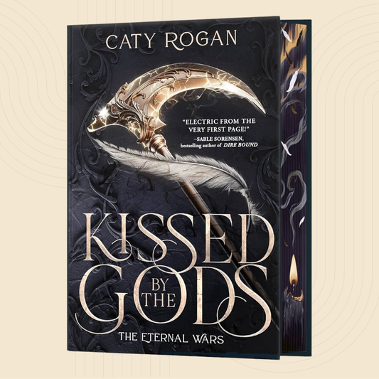 Book cover for 'Kissed by the Gods' by Caty Rogan, featuring a large ornate gold crescent moon and feather illustration on a dark black and gray background, with the title in large metallic-style lettering. A second copy of the book is partially visible behind it on the right.